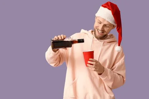 Drunk young man in Santa hat pouring drink after Christmas party on lilac bac Stock Photos