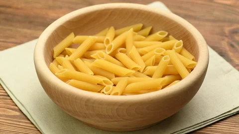Dry raw pasta penne rigate falling into a wooden bowl, slow motion Stock Footage 143866628