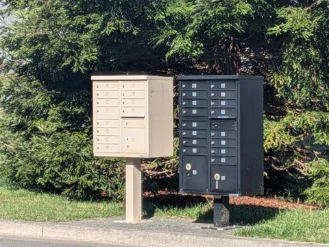 Dual community mailbox units with multiple locked slots stand beneath trees Stock Photos