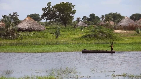 A dugout canoe moves past a village in South Sudan Stock Footage