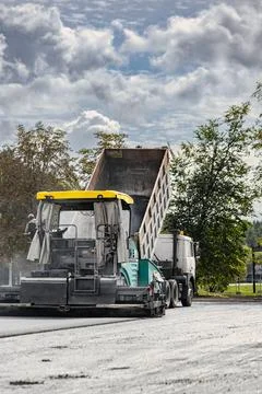 A dump truck is unloading material into a paving machine at a construction si Photos