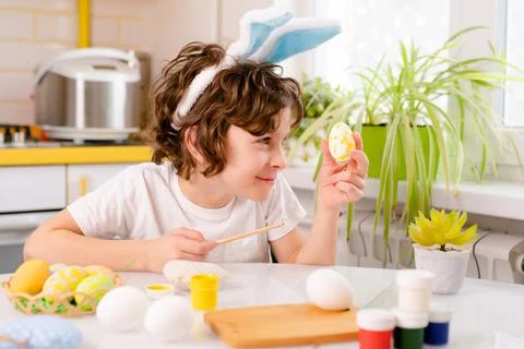 Dyeing Easter eggs. curly boy dressed in white Tshirt and blue bunny ears paints Stock Photos