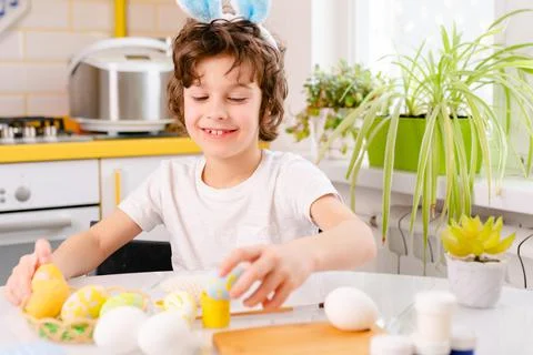 Dyeing Easter eggs. curly boy dressed in white Tshirt and blue bunny ears paints Stock Photos