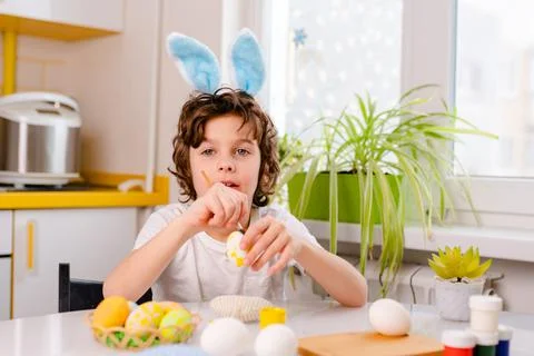 Dyeing Easter eggs. curly boy dressed in white Tshirt and blue bunny ears paints Stock Photos