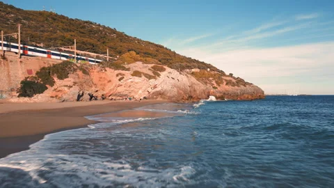 Dynamic drone shot of a beach line, blue sea, and train passing by left corner Stock Footage 223332331