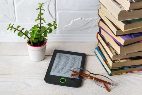E-book reader and vision glasses near stack of paper books and potted plant. Stock Photos