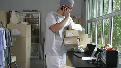 E-commerce business owner using phone and looking at laptop in store warehouse. Stock-Footage 174077547