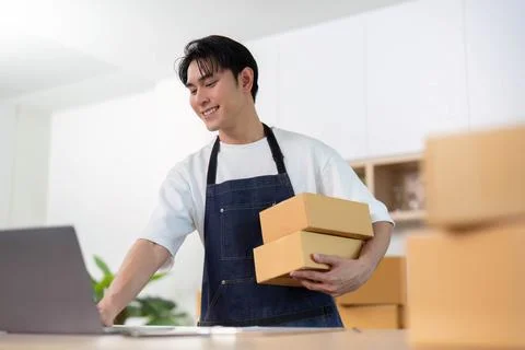 E-commerce. Smiling young man holding packages while working on laptop in a Stock Photos