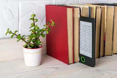 E-reader on a shelf with paper books and small potted plant. Copy space. Stock Photos