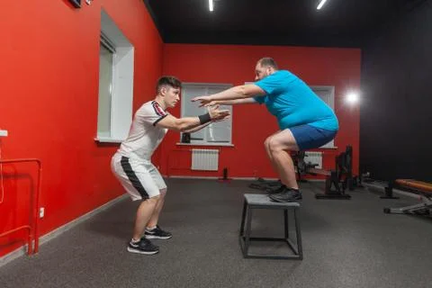Eager overweight man is doing jumping in the gym under the guidance of a Stock Photos