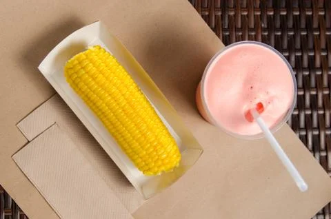 An ear of boiled corn in an eco-friendly cardboard box next to a milkshake Stock Photos