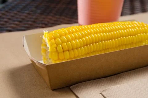An ear of boiled corn in an eco-friendly cardboard box next to a milkshake Stock Photos