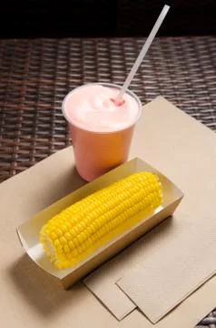 An ear of boiled corn in an eco-friendly cardboard box next to a milkshake Stock Photos