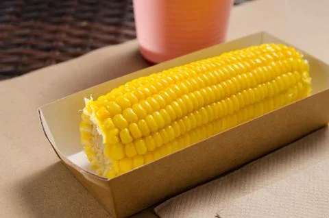 An ear of boiled corn in an eco-friendly cardboard box next to a milkshake Stock Photos