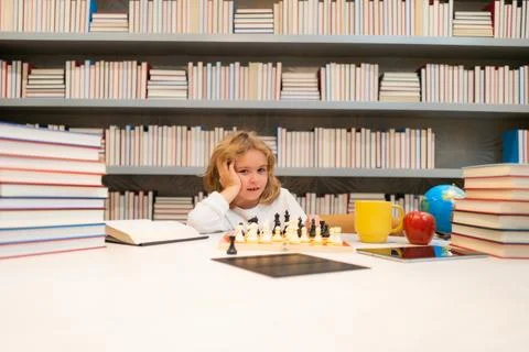 Early development. Smart child play chess in the library by the bookshelves with Fotos Stock