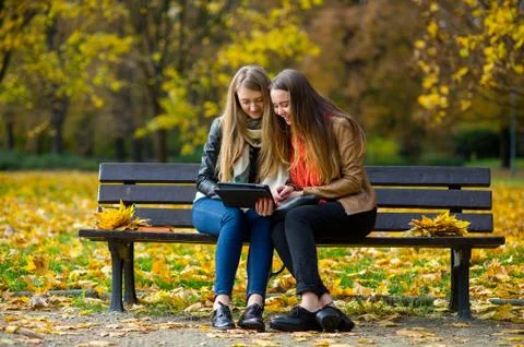 Early warm fall. Two charming girls sit on a bench in the autumn park. Stock Photos