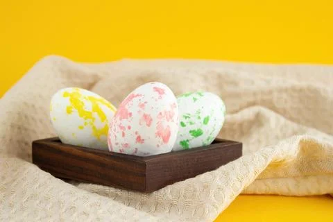 Easter-colored chicken eggs lie in a wooden low box, next to a kitchen towel  Stock Photos