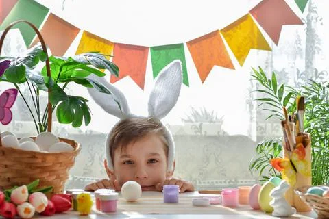 Easter composition and a boy with bunny ears looks out from behind the table. Stock Photos