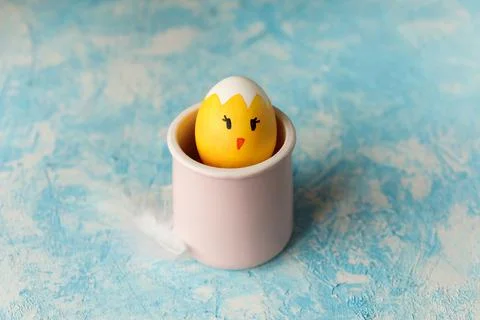 Easter egg painted as yellow chick with funny face in the pink cup on sky blue Stock Photos