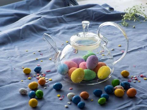 Easter eggs in glass teapot surrounded by small chocolate eggs and sprinkles Stock Photos