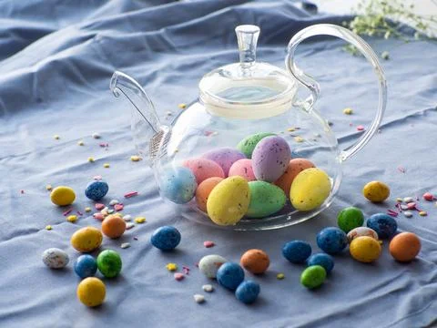 Easter eggs in glass teapot surrounded by small chocolate eggs and sprinkles Stock Photos