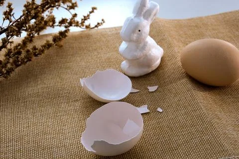 An Easter white rabbit and a broken egg on a beige tablecloth and dry branc.. Stock Photos