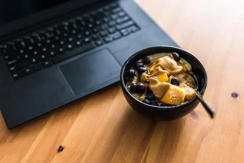 Eating mango blueberries oatmeal while working from home on a laptop, remote  Stock Photos