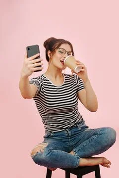 Eccentric lady taking selfie while drinking coffee. Over pink background. Stock Photos