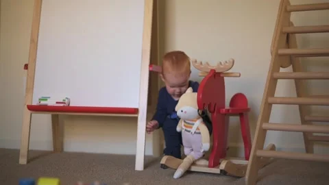 Ed-haired toddler cuddles soft toy next to wooden rocking horse in cozy playroom Stockbeeldmateriaal 295839118