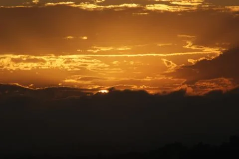 Edge of sun behind clouds and mountain range silhouette with orange, cloudy sky Stock Photos