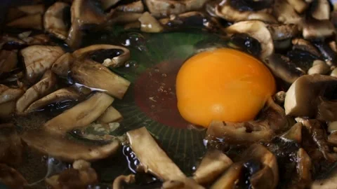 Egg adding to omelette with mushrooms on black frying pan close up Stock Footage 297478182