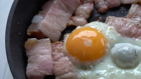 Egg and bacon are fried in a pan. Appetizing Pieces of bacon and eggs in a hot Stock Footage 144431635