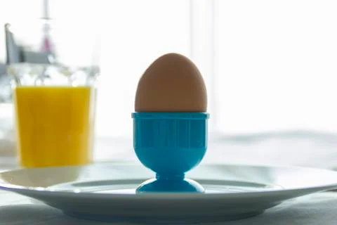 An egg in a blue plastic egg cup on a plate during breakfast in front of a wi Stock Photos
