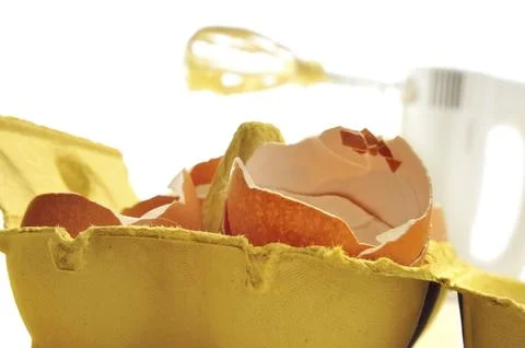 Egg carton with freshly cracked eggshells and mixer in the background Stock Photos