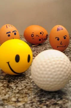 Eggs with drawn faces, smiling yellow ball, and golf ball on granite countert Stock Photos
