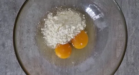Eggs with flour in a bowl. Flour is poured from a spoon into a plate with eggs. Stock Footage 220105437