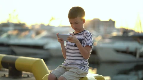 An eight-year-old boy sits on the shore and plays the phone against the backdrop Stock Footage 263699788