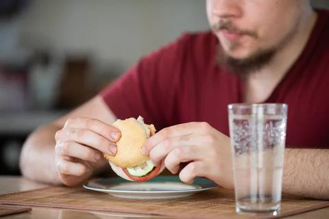 Ein bärtiger Mann isst ein Sandwich am Tisch. A man sits at a table. In fr.. Stock Photos