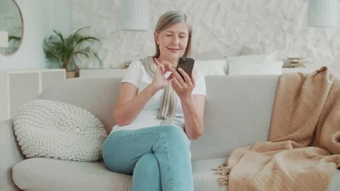 Elder middle age woman using smartphone sitting on sofa smiling at home in Vídeos de archivo 224539340