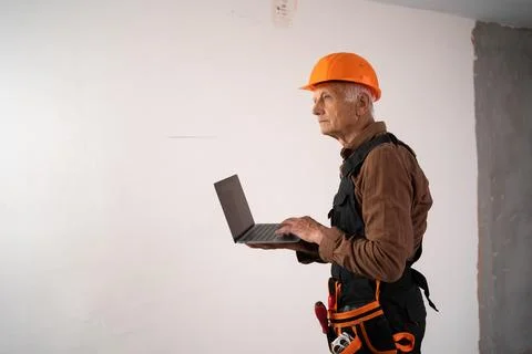 Elderly builder in safety helmet using laptop at construction site preparin.. Stock Photos