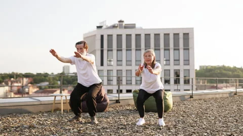 Elderly couple doing squats during morning warm-up on urban rooftop Stock Footage 309907195