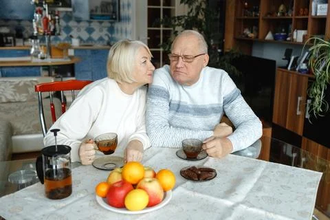 An elderly couple sits at the table, drinks tea and kisses. Stock Photos