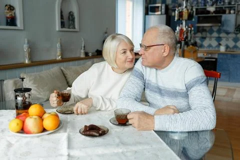 An elderly couple sits at the table, drinks tea and kisses. Stock Photos