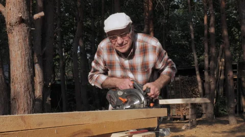 Elderly craftsman skillfully using a power saw to shape wood in a serene forest Stock Footage 285172658
