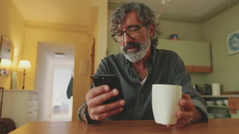 Elderly disappointed man drinks coffee from mug and uses mobile phone at home Video stock 229505780