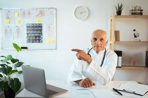 Elderly doctor looking at camera pointing with finger at advertising space si Stock Photos
