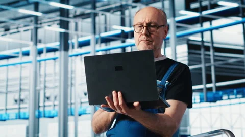 Elderly engineer uses laptop in smart factory to test automated systems Stock Footage 305481623