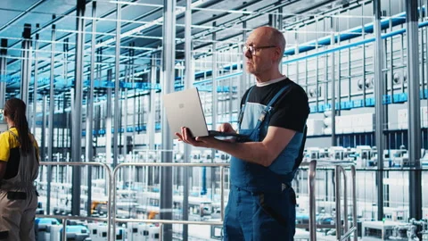 Elderly engineer uses laptop in smart factory to test automated systems Stock Footage 305501617