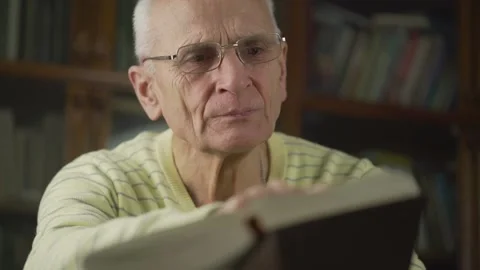 Elderly gray haired man with glasses is attentively reading large book. Stock Footage 148283200