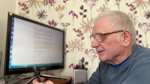 An elderly gray-haired man with glasses sits in front of a monitor, talking on a Stock Footage 233570448